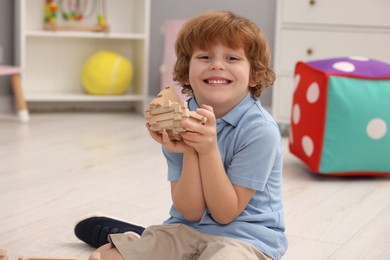 Happy little boy playing with piece of wooden construction set on floor in room. Child's toy