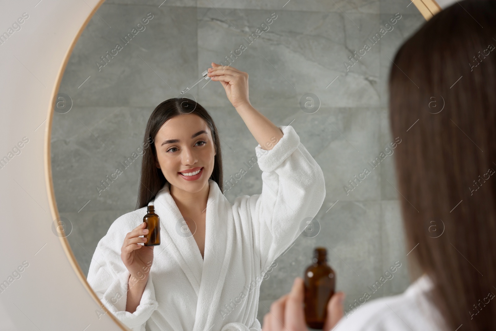 Photo of Happy young woman applying essential oil onto hair roots near mirror in bathroom