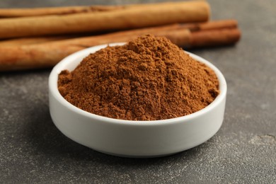 Photo of Bowl of cinnamon powder and sticks on grey table, closeup