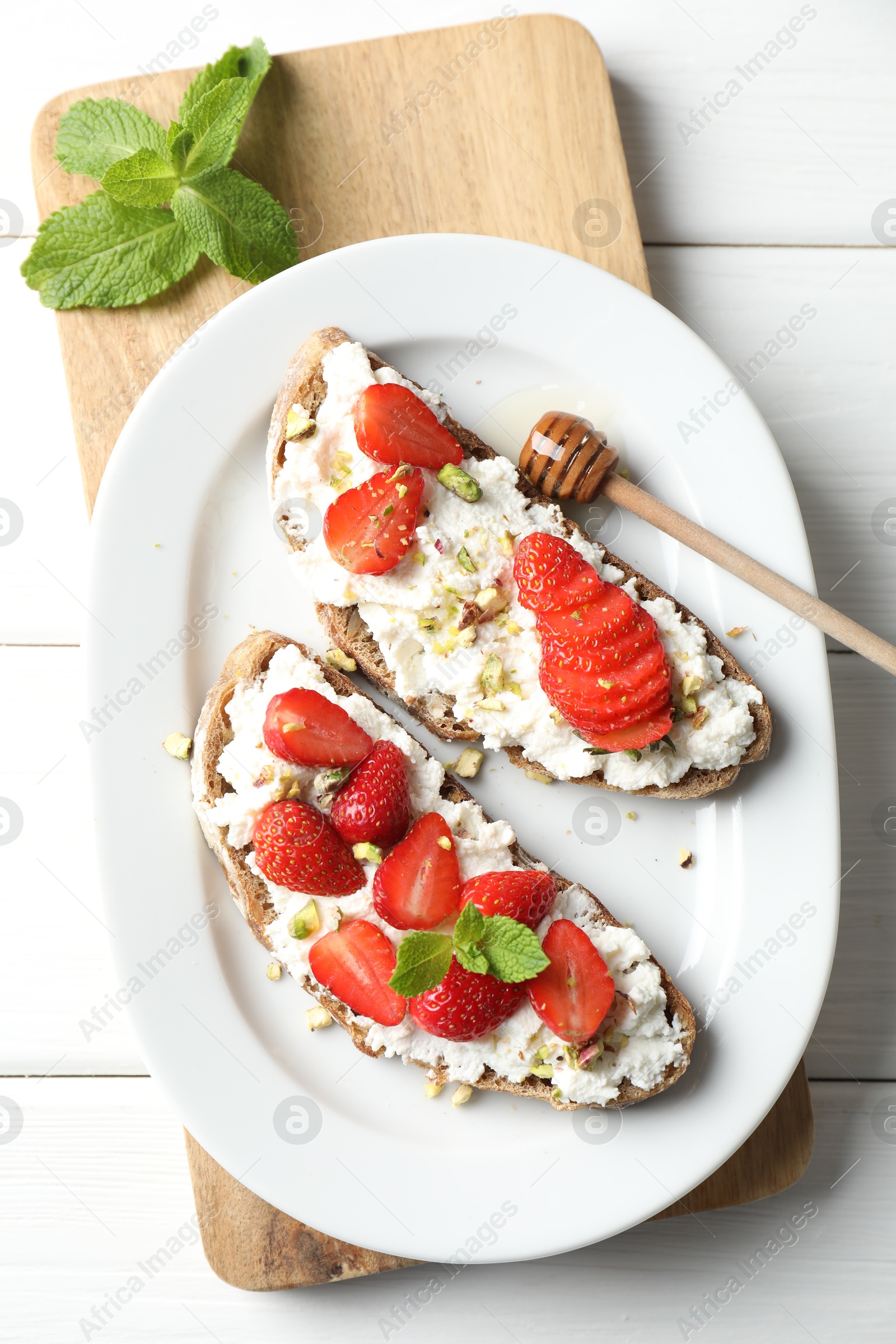 Photo of Delicious ricotta bruschettas with strawberry, mint and pistachios served with honey on white wooden table, flat lay