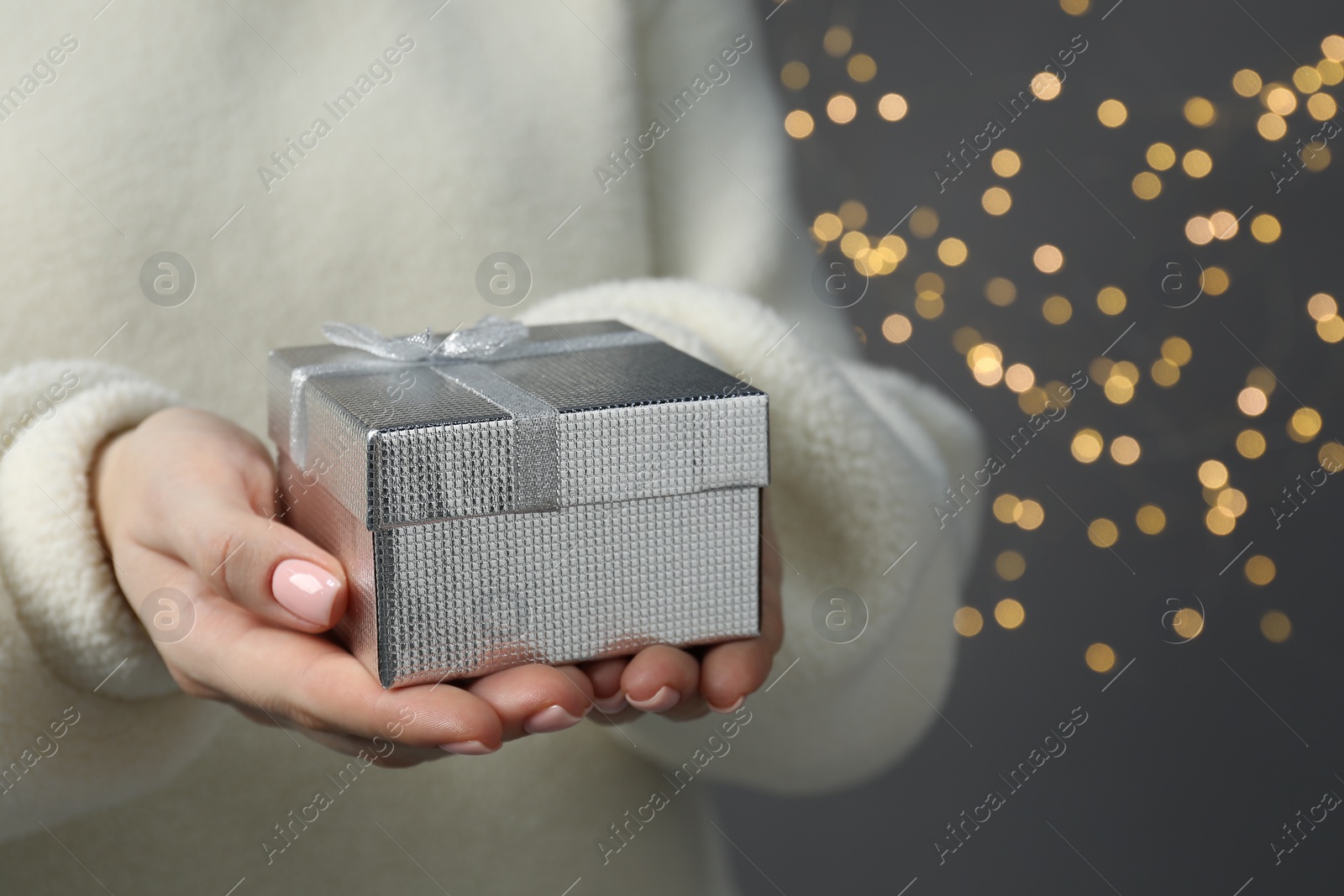 Photo of Christmas present. Woman holding gift box against grey background with blurred lights, closeup. Space for text