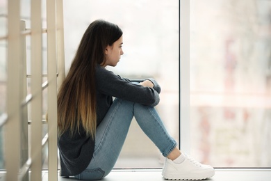 Upset teenage girl sitting at window indoors. Space for text