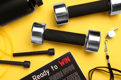 Metal dumbbells, whistle, magazine and skipping rope on yellow background, flat lay