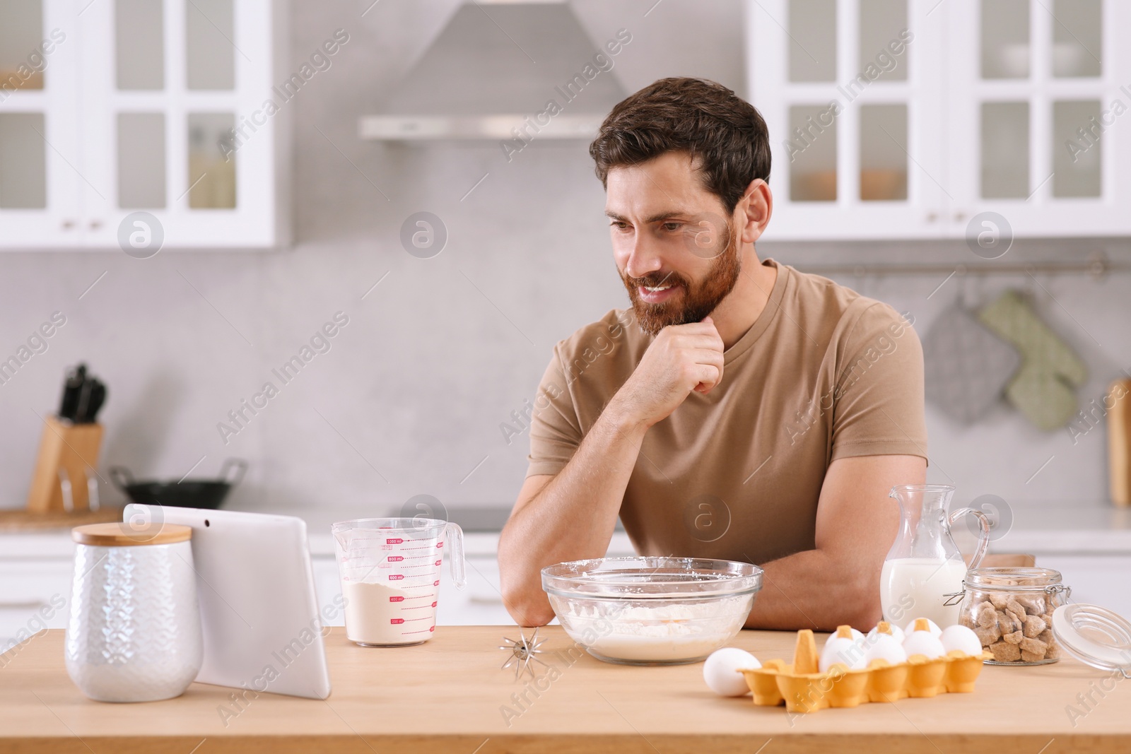 Photo of Man making dough while watching online cooking course via tablet in kitchen