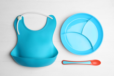 Photo of Colorful plastic dishware and silicone bib on white wooden table, flat lay. Serving baby food