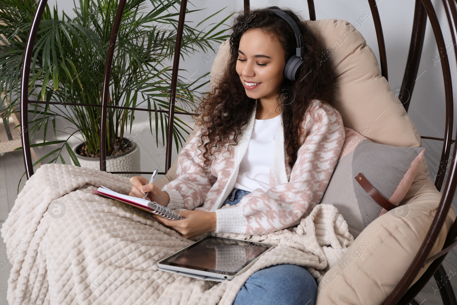 Photo of African American woman with headphones and tablet studying in egg chair at home. Distance learning