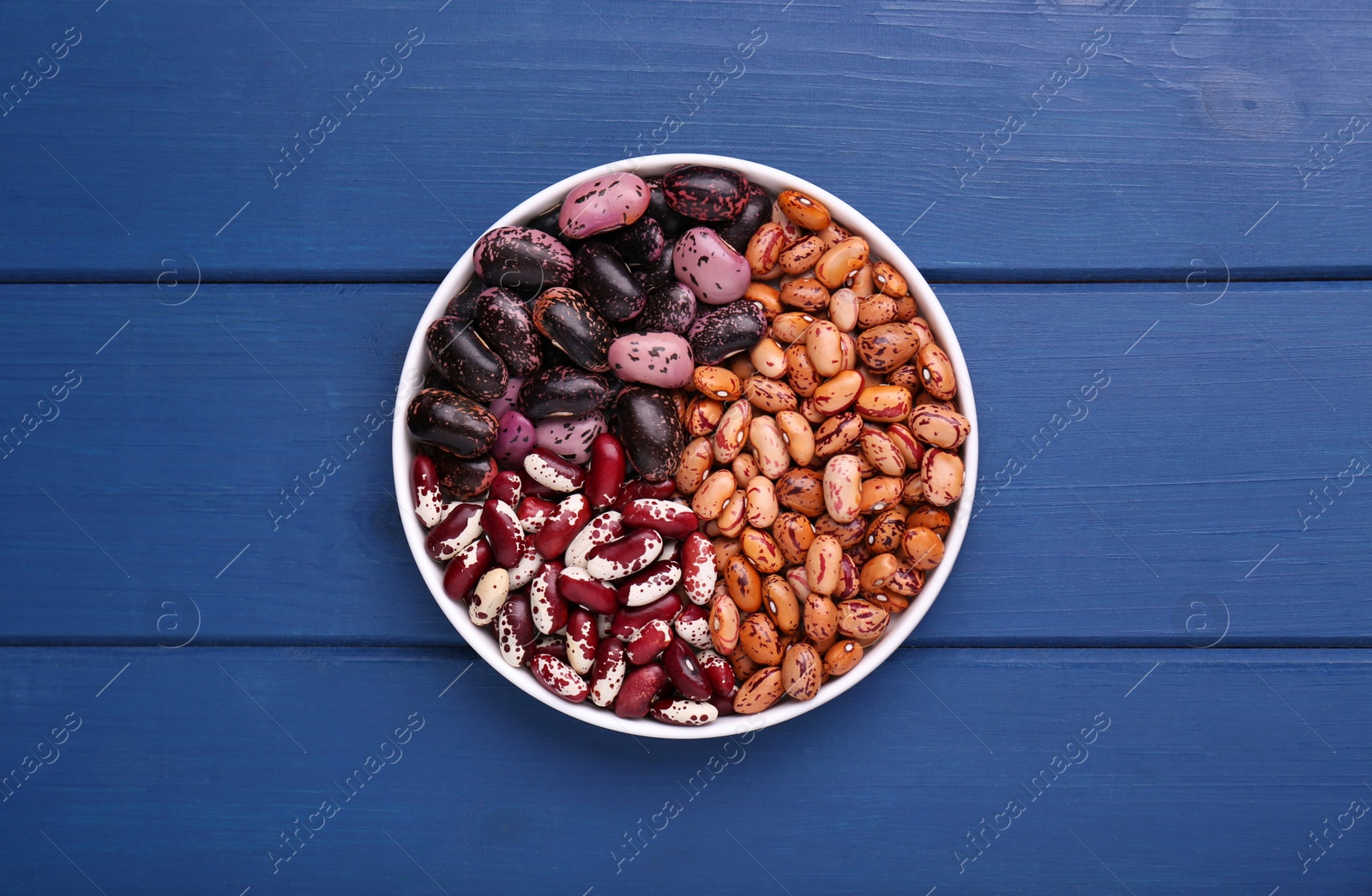 Photo of Different kinds of dry kidney beans in bowl on blue wooden table, top view