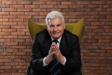 Happy mature businessman sitting in armchair near brick wall