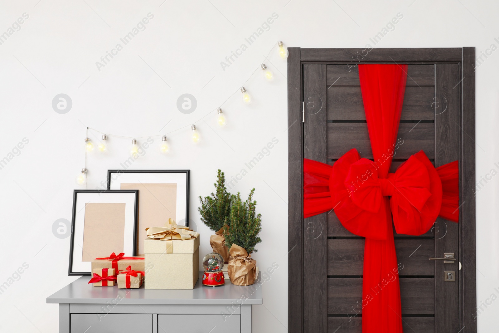 Photo of Wooden door with beautiful bow near chest of drawers with empty frames and gift boxes. Christmas decoration