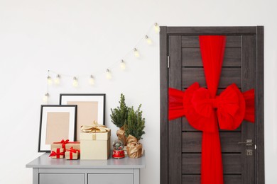 Photo of Wooden door with beautiful bow near chest of drawers with empty frames and gift boxes. Christmas decoration