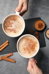 Photo of Women holding glass cups of delicious eggnog with cinnamon and anise at grey table, top view