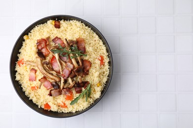 Tasty couscous with mushrooms and bacon in bowl on white tiled table, top view. Space for text