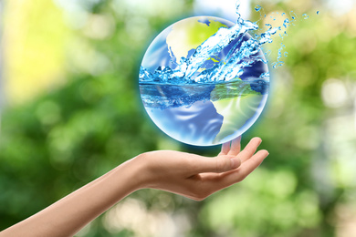 Image of Woman holding icon of Earth with water splashes on blurred green background, closeup. Ecology concept