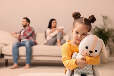 Photo of Sad little girl with toy and her arguing parents on sofa in living room
