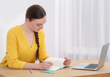 Woman writing in notebook while working on laptop at wooden table indoors