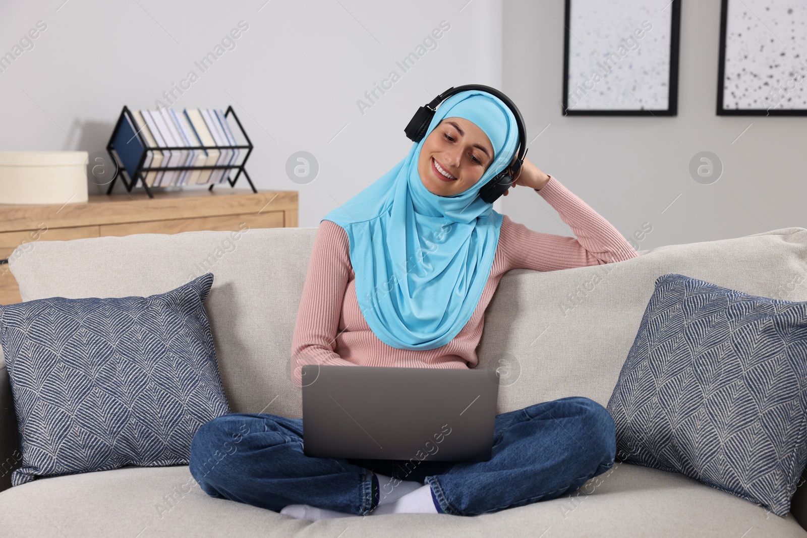 Photo of Muslim woman in headphones using laptop at couch in room