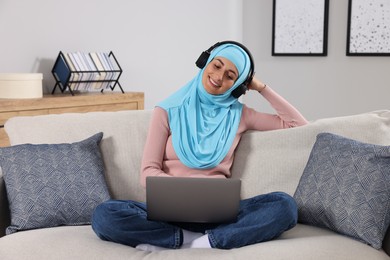 Muslim woman in headphones using laptop at couch in room