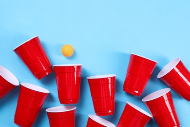 Plastic cups and ball on light blue background, flat lay. Beer pong game