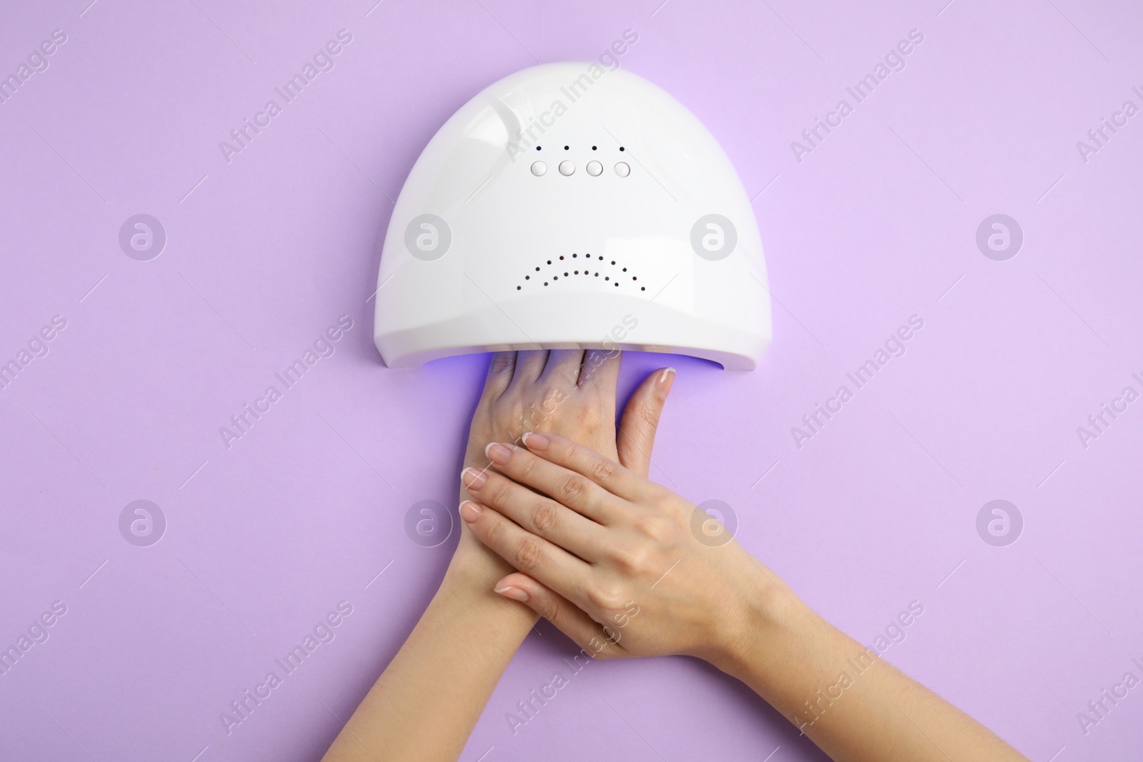 Photo of Woman using ultraviolet lamp to dry gel nail polish on violet background, top view