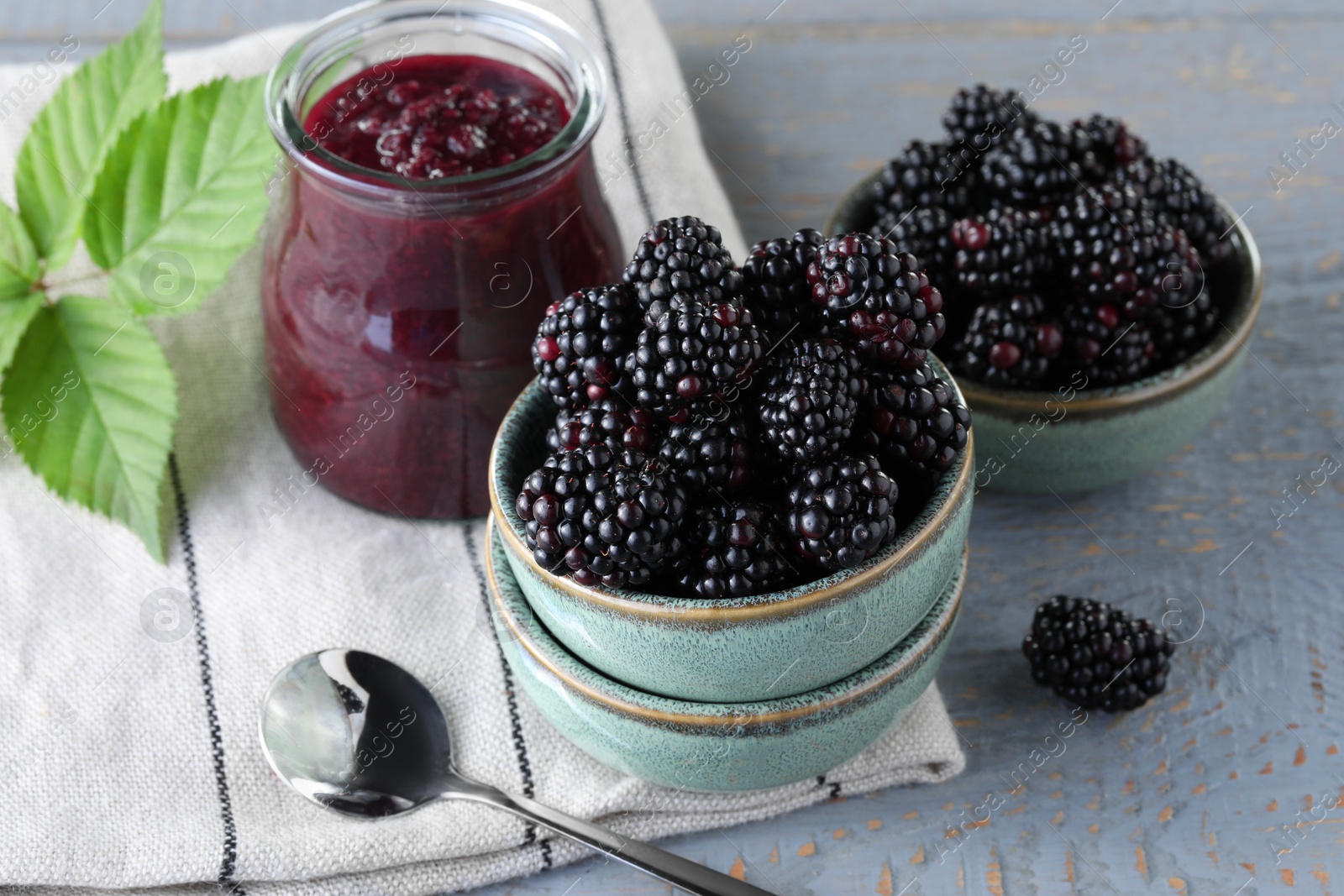 Photo of Fresh ripe blackberries, tasty jam and leaves on light grey wooden table