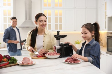Photo of Happy family making dinner together in kitchen, little girl using modern meat grinder while her mother cutting onion