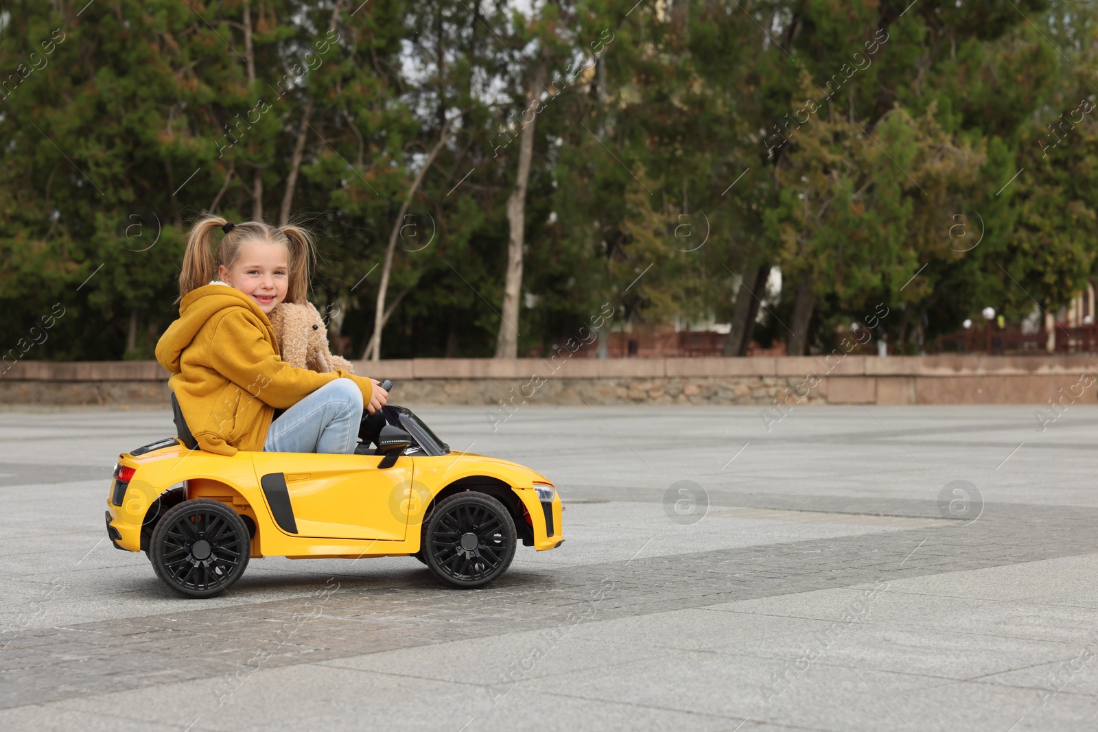 Photo of Cute little girl with toy bunny driving children's car on city street. Space for text