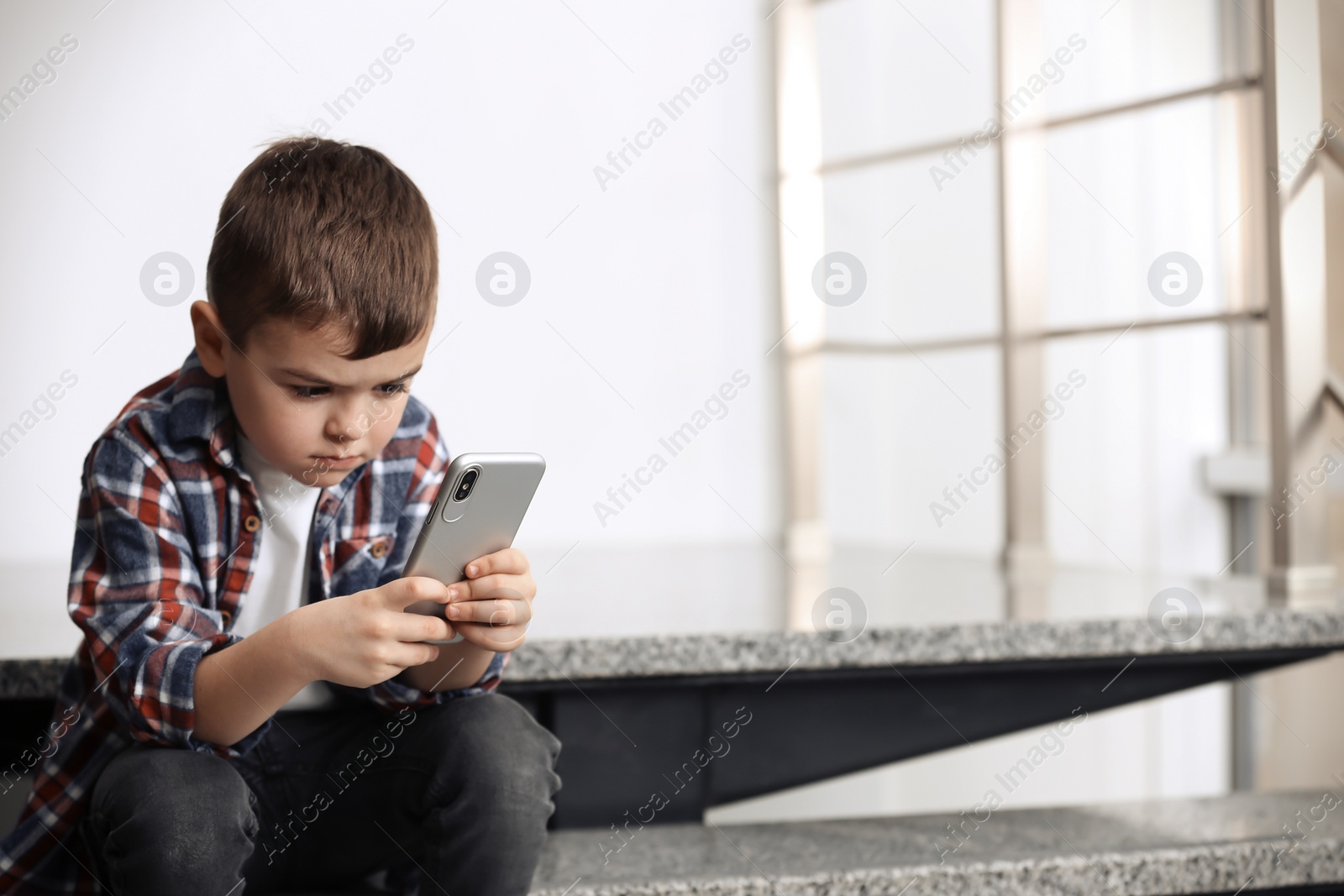 Photo of Sad little boy with mobile phone sitting on stairs indoors