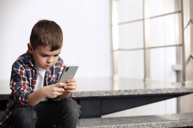 Sad little boy with mobile phone sitting on stairs indoors