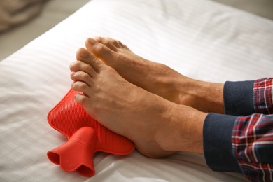 Photo of Man warming feet with hot water bottle on bed, closeup