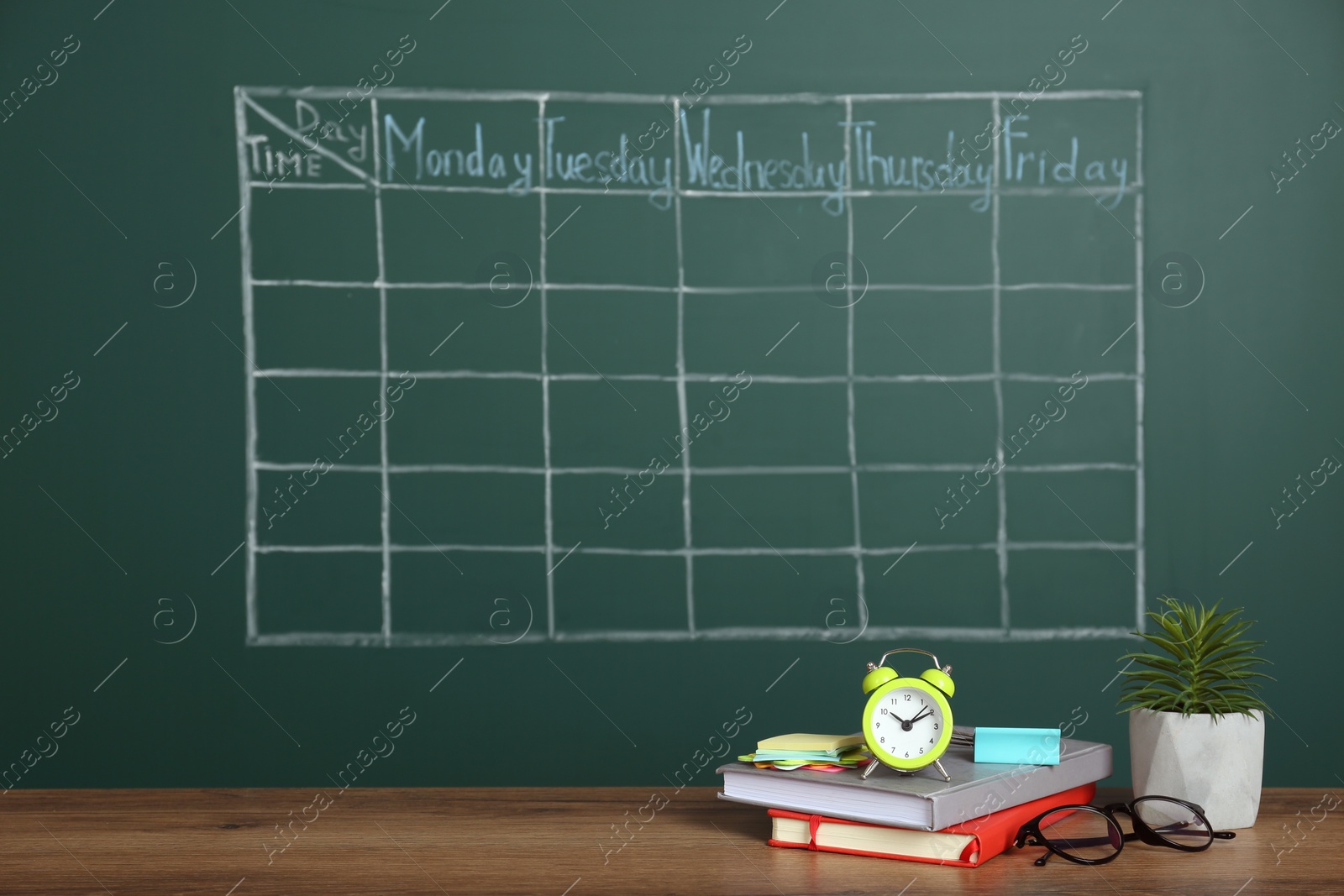 Photo of Alarm clock, stationery and plant on wooden table near green chalkboard with drawn school timetable
