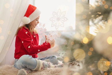 Photo of Little girl in Santa hat decorating window with snowflakes at home