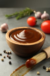 Tasty barbeque sauce in bowl, brush and peppercorns on grey table, closeup