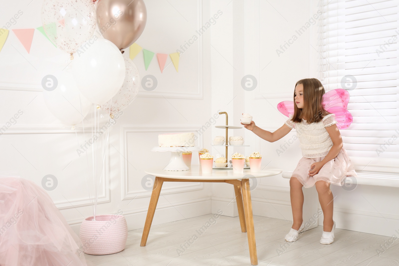 Photo of Cute little girl wearing fairy costume at table with desserts in decorated room