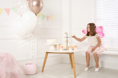 Photo of Cute little girl wearing fairy costume at table with desserts in decorated room