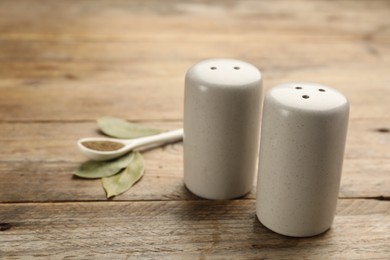 Photo of Ceramic salt and pepper shakers, bay leaves with spoon on wooden table, closeup