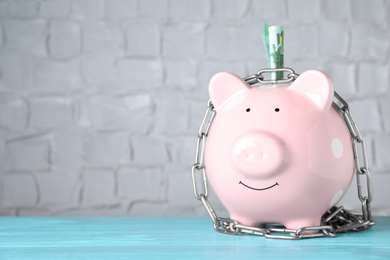 Piggy bank with steel chain and banknote on light blue wooden table, space for text. Money safety concept