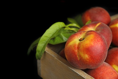 Wooden crate with tasty peaches and leaves on black background, closeup