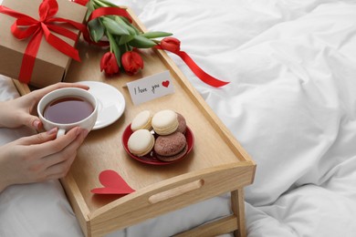 Photo of Tasty breakfast served in bed. Woman with tea, macarons, gift box, flowers and I Love You card at home, closeup