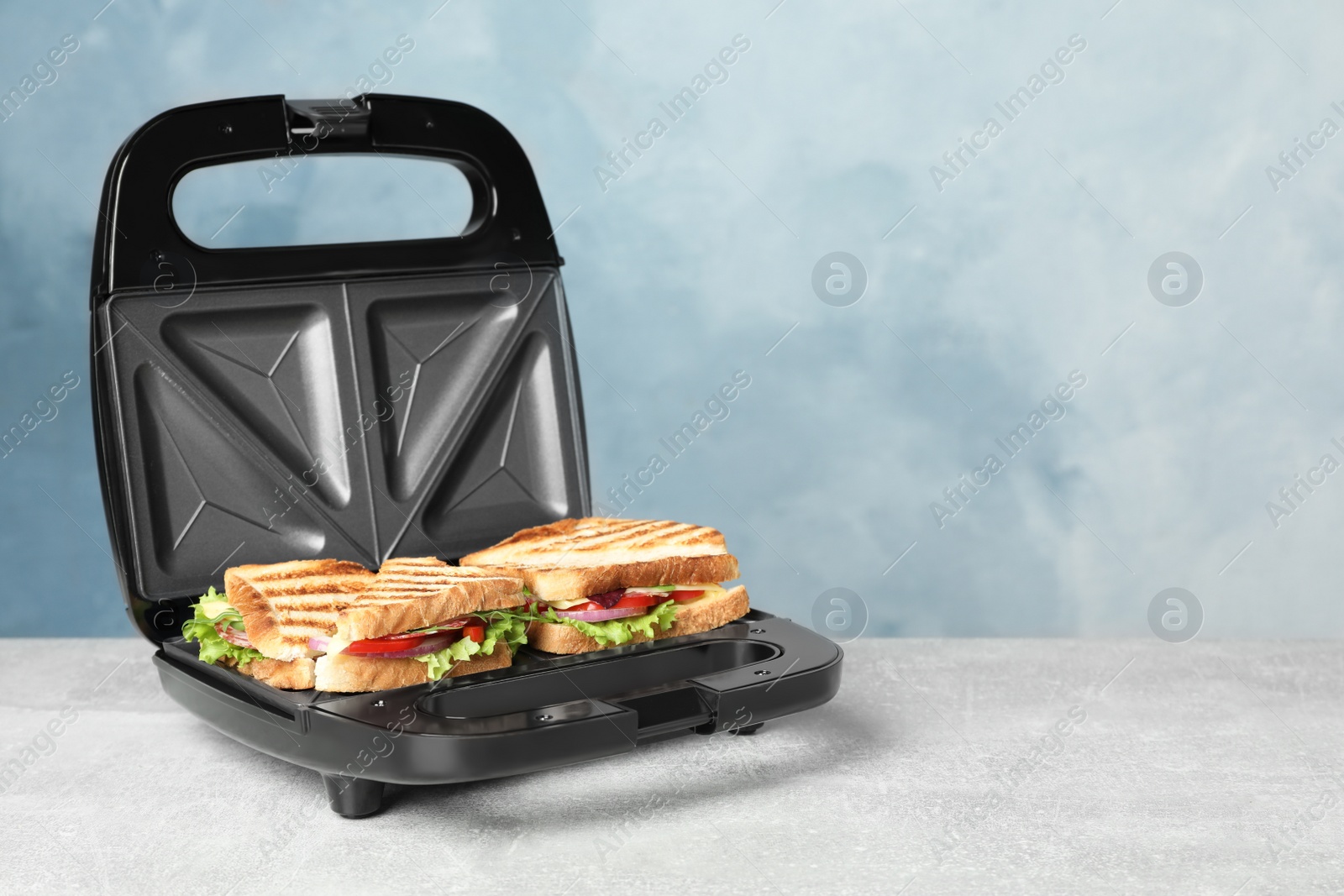 Photo of Modern grill maker with sandwiches on grey table near light blue wall, space for text