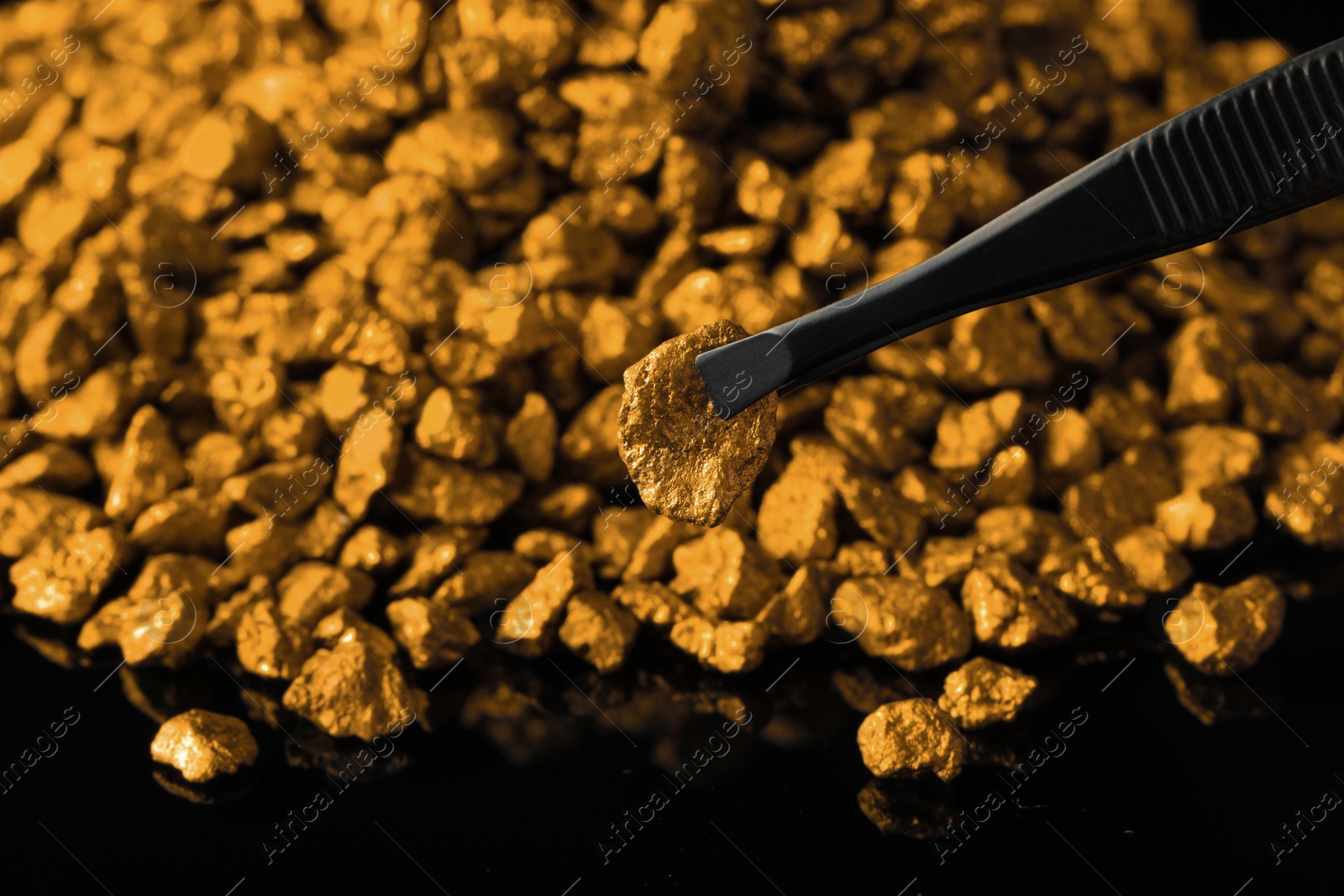 Photo of Tweezers with gold nugget above pile on black background, closeup