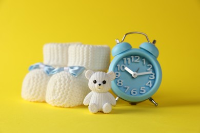 Alarm clock, toy bear and baby booties on yellow background. Time to give birth