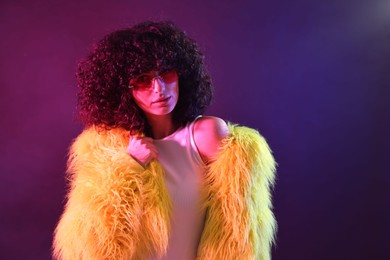 Beautiful young woman in yellow fur coat and sunglasses on color background in neon lights