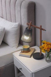 Stylish lamp, book, hot drink and flowers on bedside table indoors. Bedroom interior element