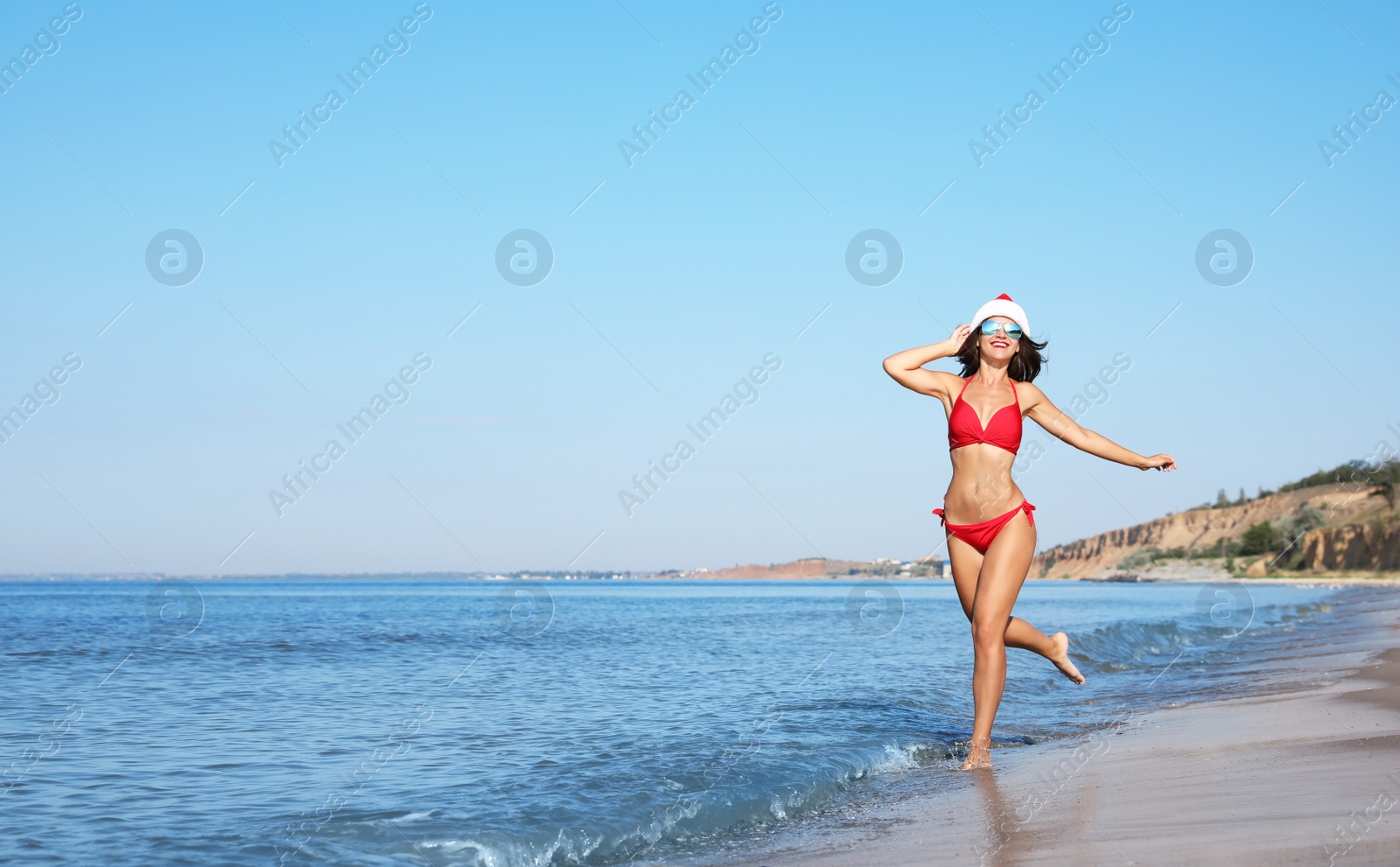 Photo of Young woman wearing Santa hat and bikini on beach, space for text. Christmas vacation