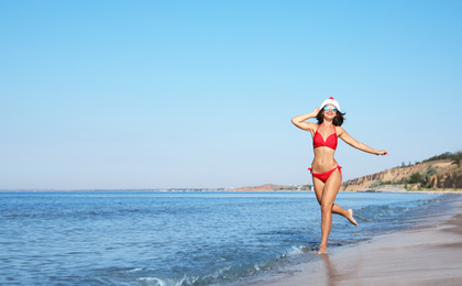 Photo of Young woman wearing Santa hat and bikini on beach, space for text. Christmas vacation