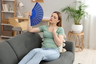 Photo of Woman waving blue hand fan to cool herself on sofa at home
