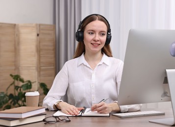 Photo of E-learning. Young woman taking notes during online lesson at wooden table indoors