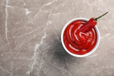 Bowl with spicy chili sauce and fresh pepper on gray table, top view