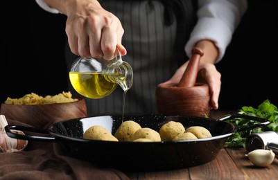 Woman pouring oil onto frying pan with raw falafel balls at wooden table, closeup