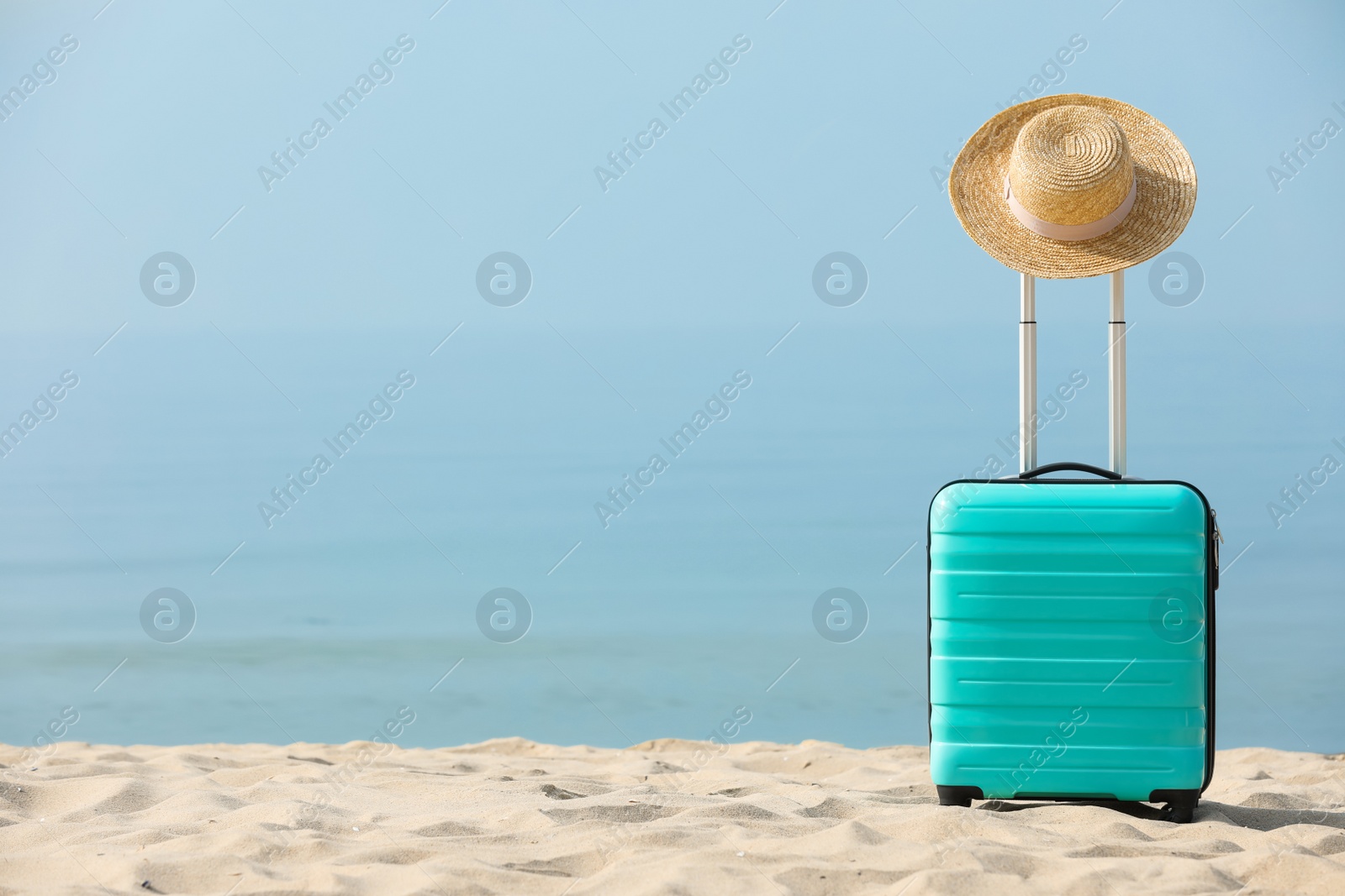 Photo of Turquoise suitcase with straw hat on sandy beach, space for text